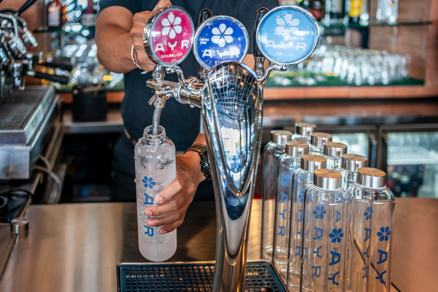 Bartender filling a reusable AYR Water glass bottle with purified chilled water from an on-site still, chilled and sparkling water dispensing system.